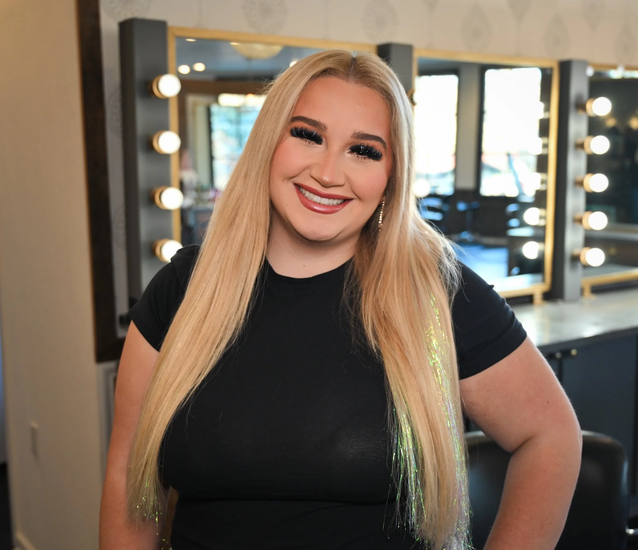 A woman with long blonde hair, wearing a black top, smiling in front of a dressing room mirror with lights.