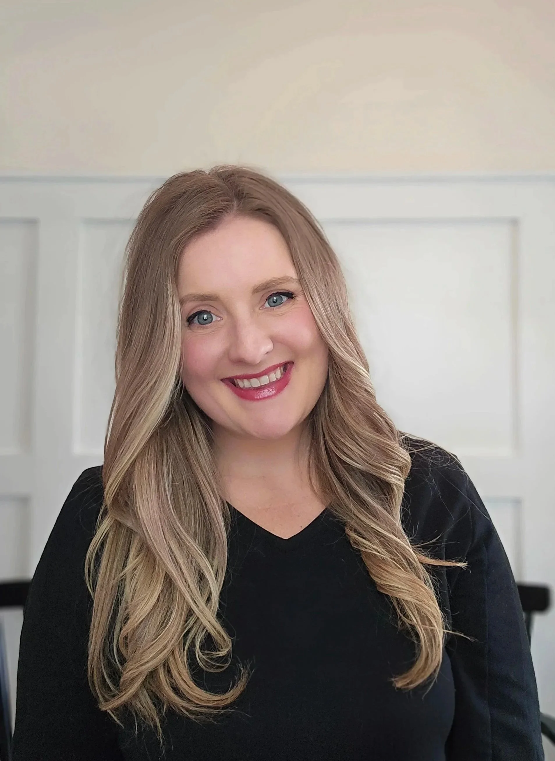 A woman with long wavy blonde hair wearing a black top, smiling, against a white paneled wall background.