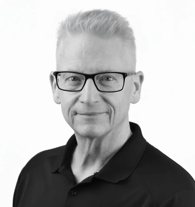Black and white portrait of a middle-aged man with short gray hair, wearing glasses and a black polo shirt, smiling gently.