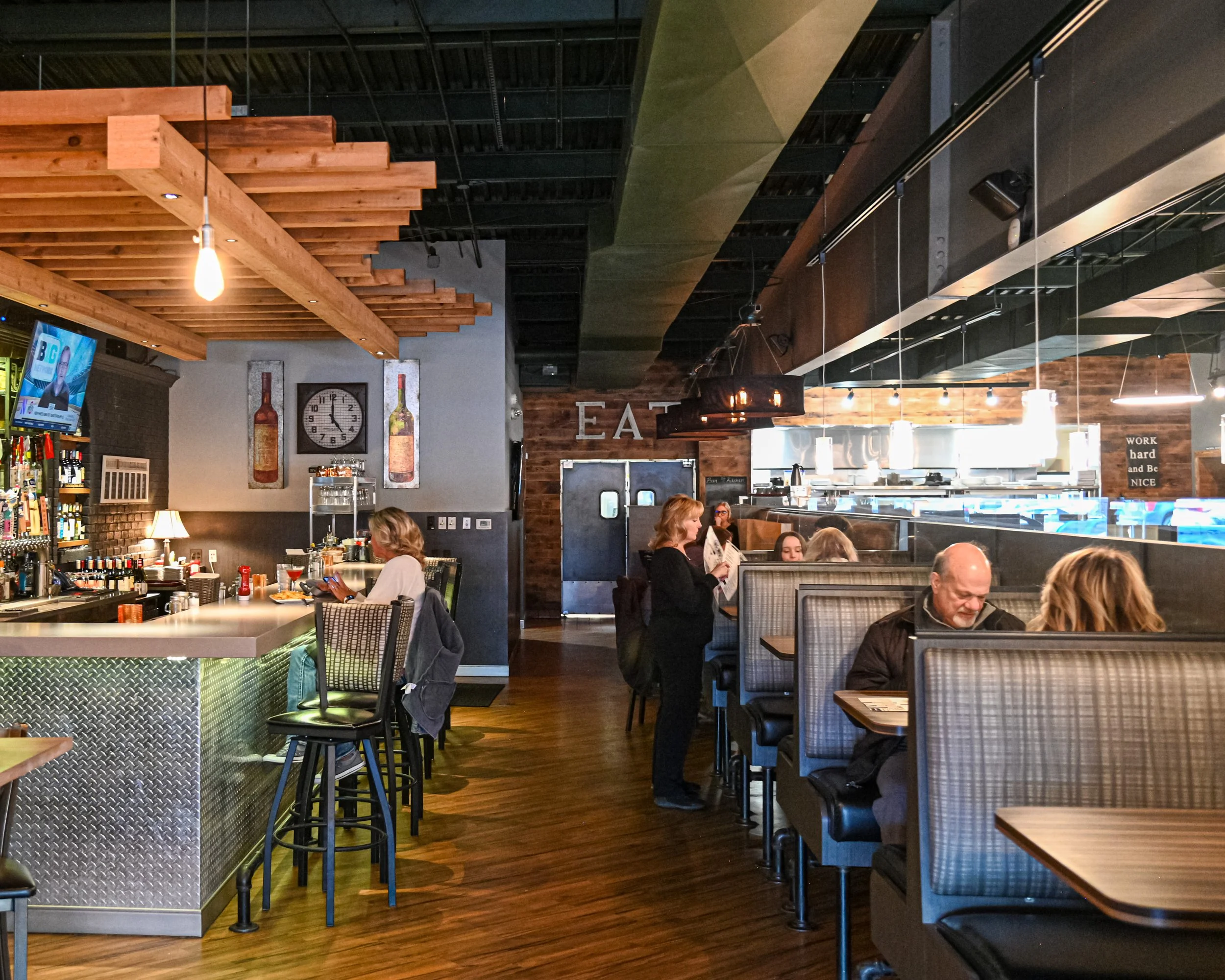 Inside a casual restaurant or cafe with wooden and metal decor, several people are sitting at tables, some looking at menus or using phones, and a television is mounted on the wall.