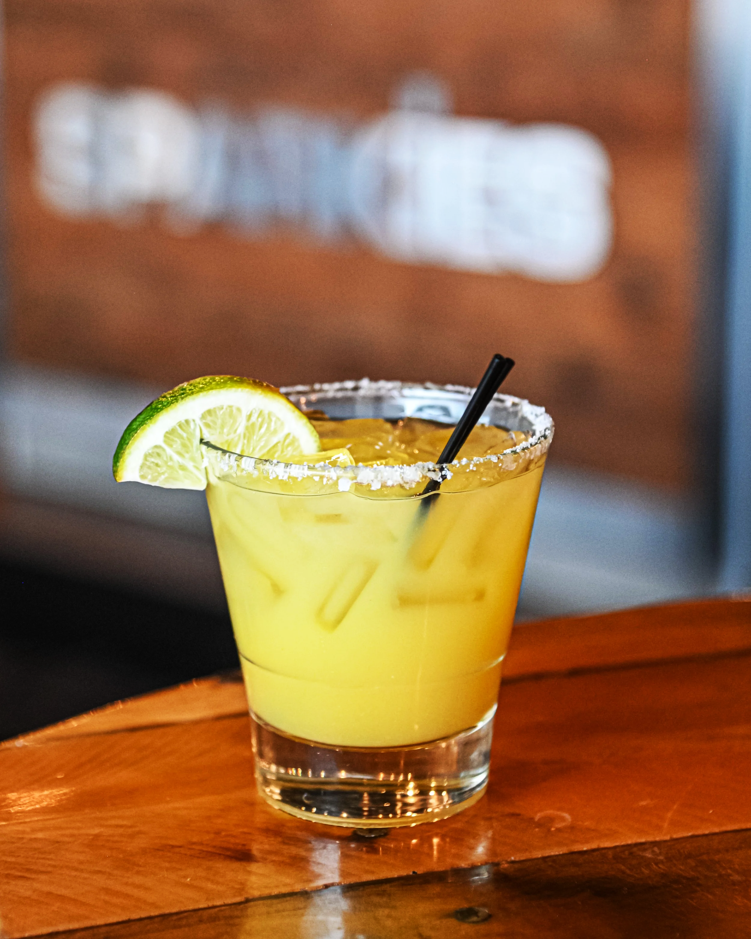 A margarita cocktail in a glass with salted rim, lime wedge, and a black straw on a wooden table.
