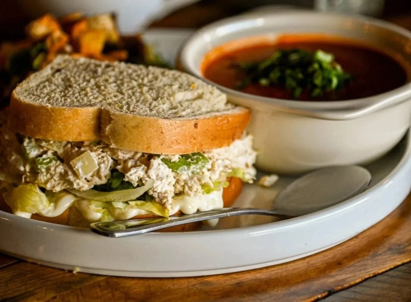 A plate of chicken salad sandwich with two slices of whole wheat bread, coleslaw filling, and a side of tomato-based soup garnished with herbs.