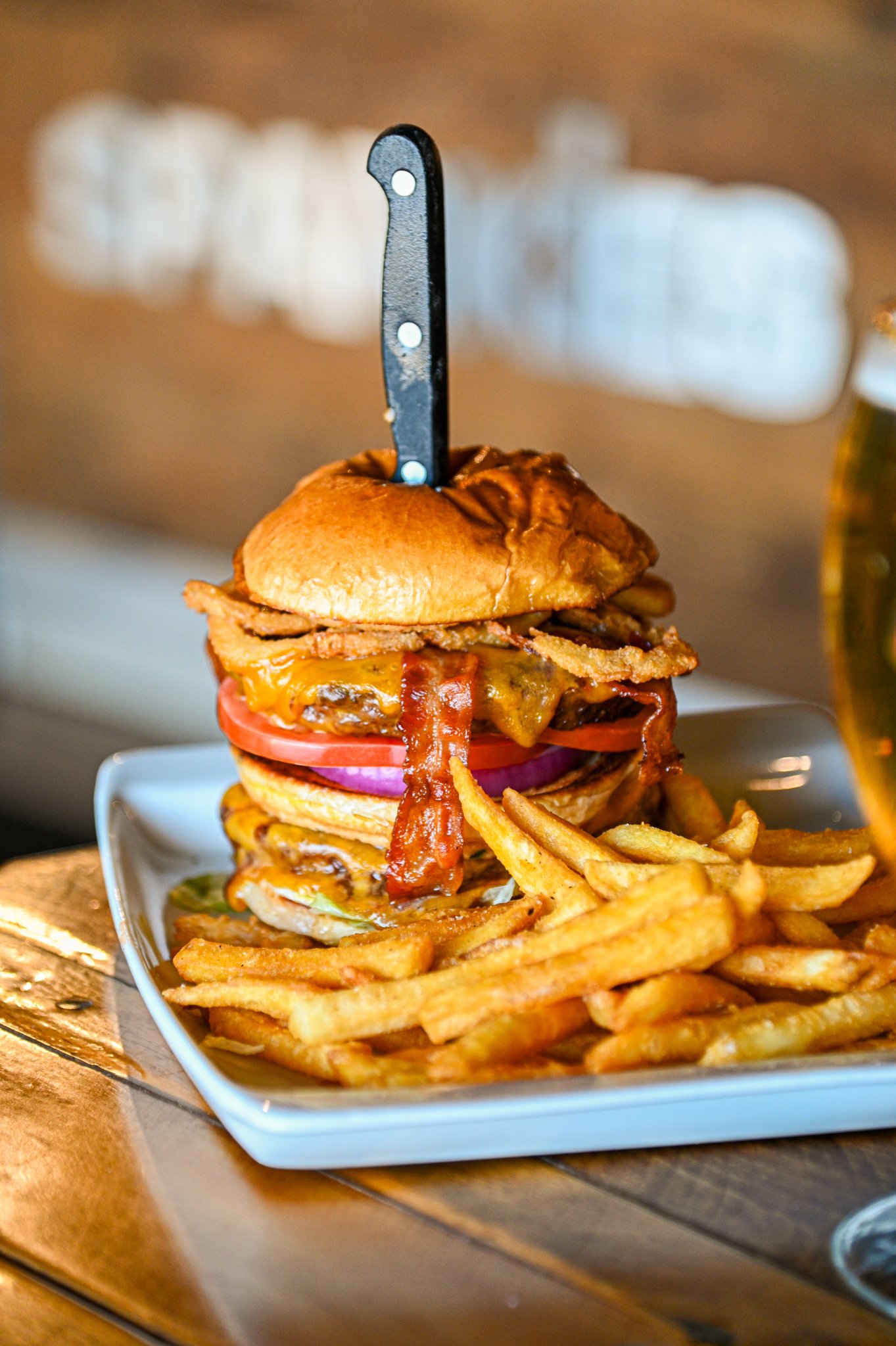 Close-up of a large cheeseburger with bacon, tomato, onions, and lettuce topped with a bun, served with French fries on a white dish.