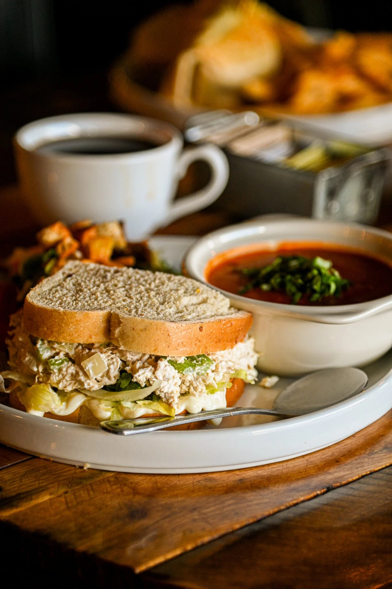 A sandwich with chicken salad, lettuce, tomato, and sliced bread on a white plate, with a cup of coffee and a bowl of sauce in the background.