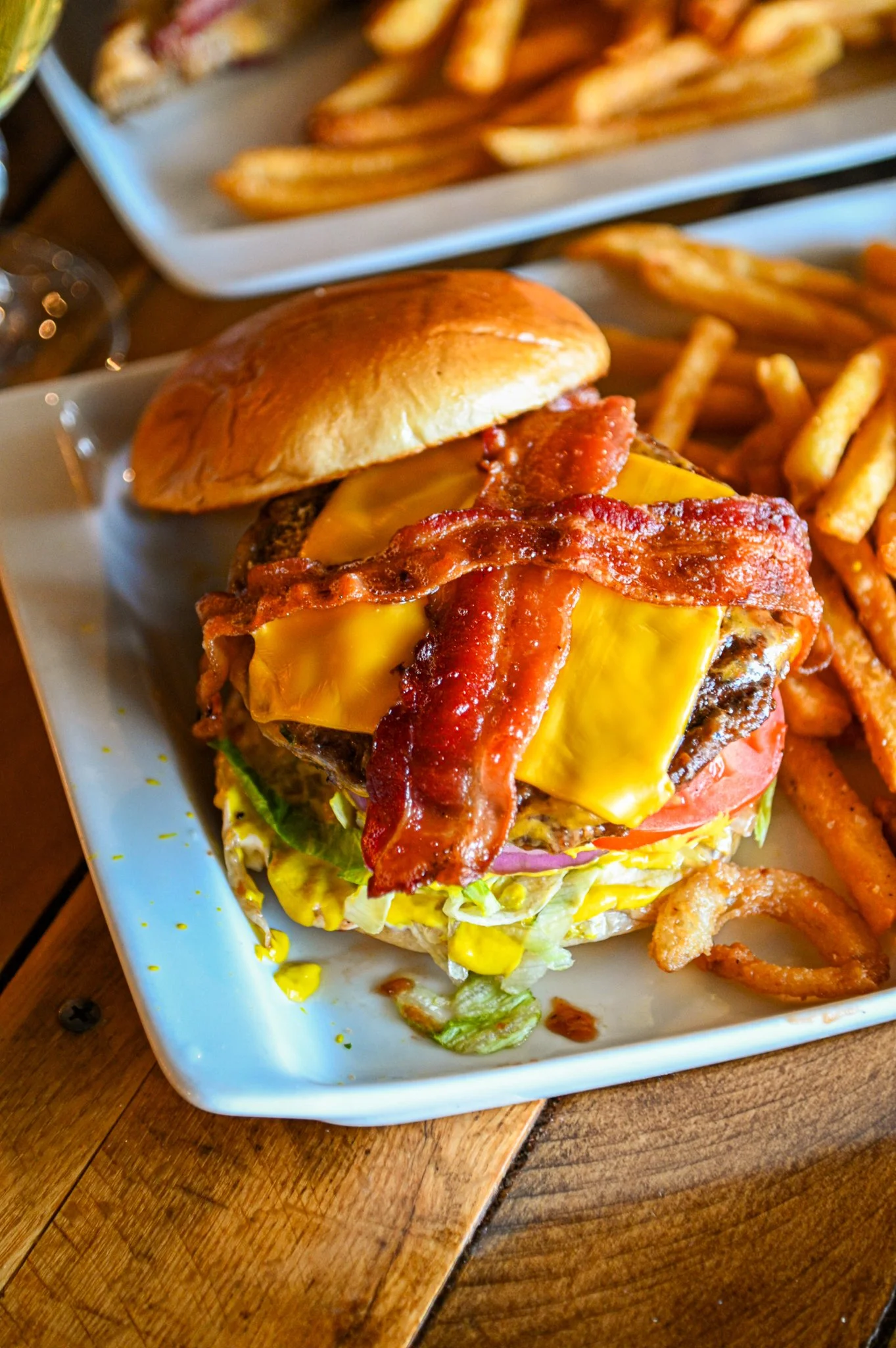 A plate with a cheeseburger topped with bacon, lettuce, tomato, and cheddar cheese, accompanied by French fries and onion rings on a wooden table.