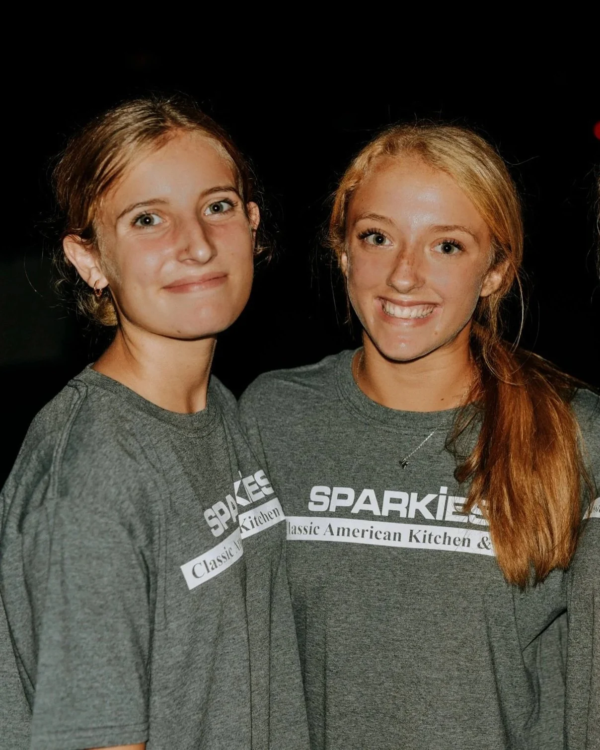 Two young women with red hair and fair skin standing close together, smiling, wearing gray t-shirts with 'SPARKIES' and 'Classic American Kitchen' printed on them, against a dark background.