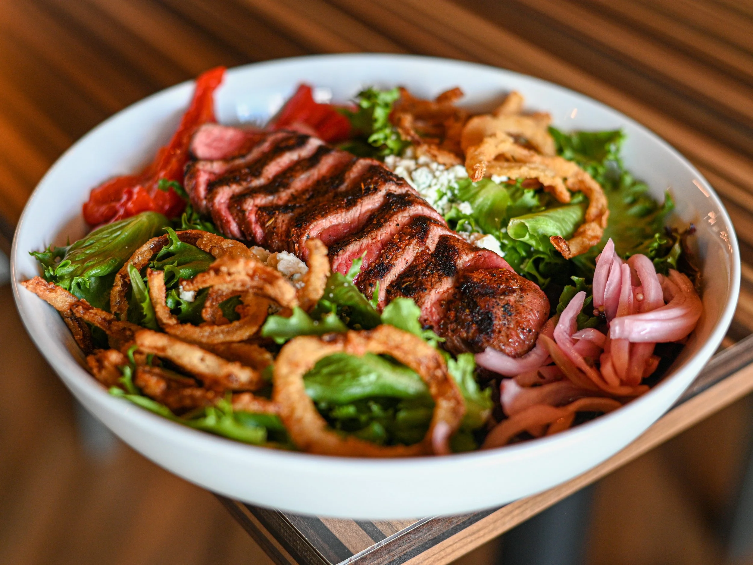 A white bowl containing a sliced grilled steak, mixed greens, fried onion strings, cherry tomatoes, and shredded cheese on a wooden table.