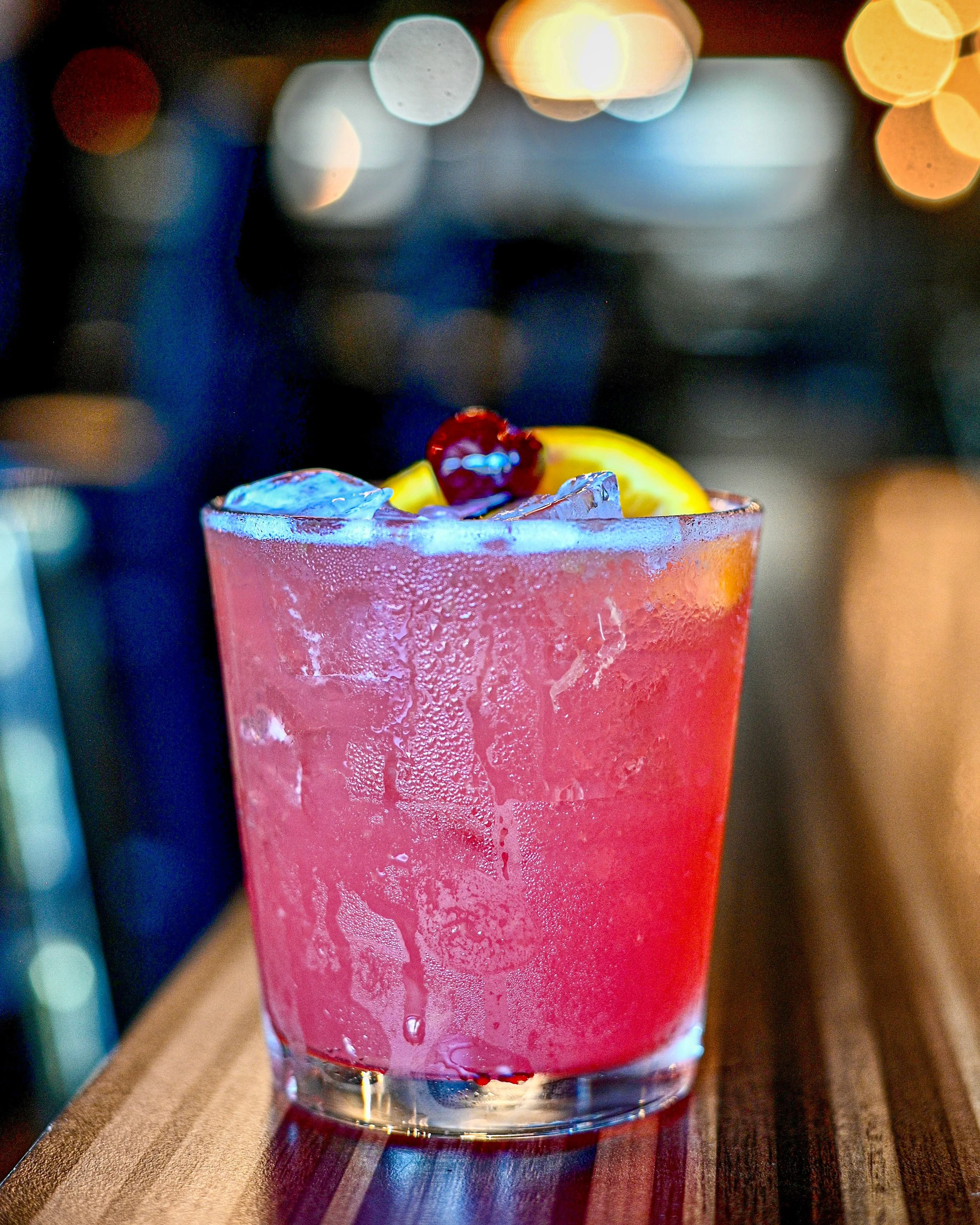 Close-up of a pink cocktail glass filled with ice and garnished with a cherry and orange slices, set on a wooden surface.