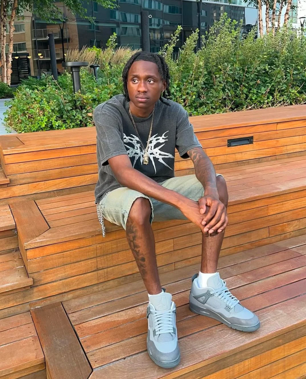A young man with dreadlocks sitting on a wooden bench outdoors with greenery and modern buildings in the background, wearing a graphic T-shirt, beige shorts, and gray sneakers.