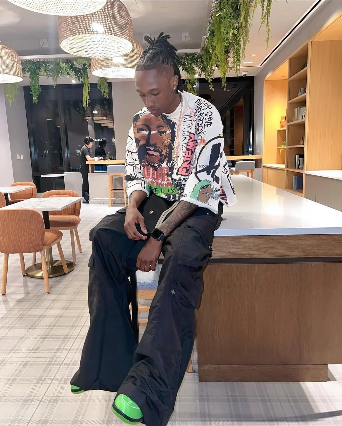 Young man with dreadlocks sitting on a chair at a modern, well-lit cafe with wooden furniture and hanging plants, looking down.