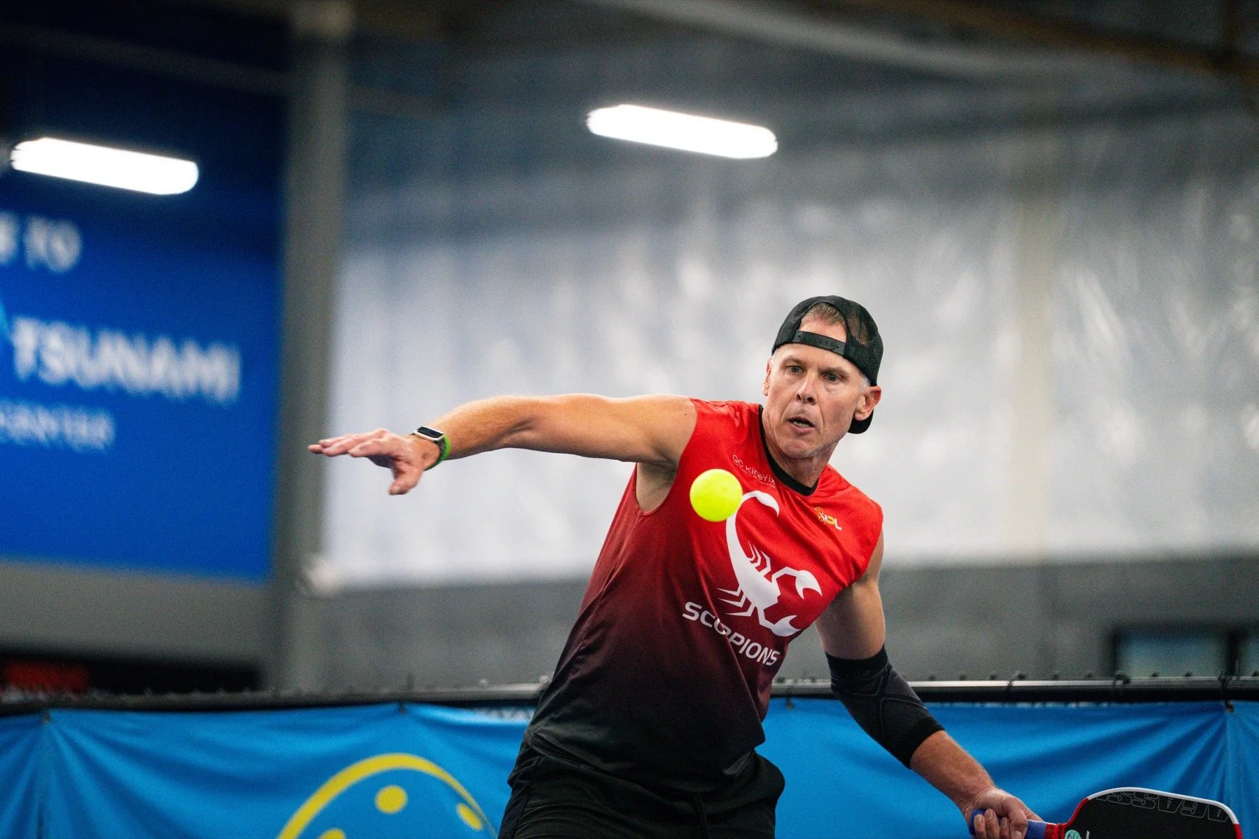 A man wearing a black cap backwards, red sleeveless shirt, and black arm sleeves, playing pickleball indoors. He is about to hit a yellow ball with a paddle, with a focus on his action and concentration.