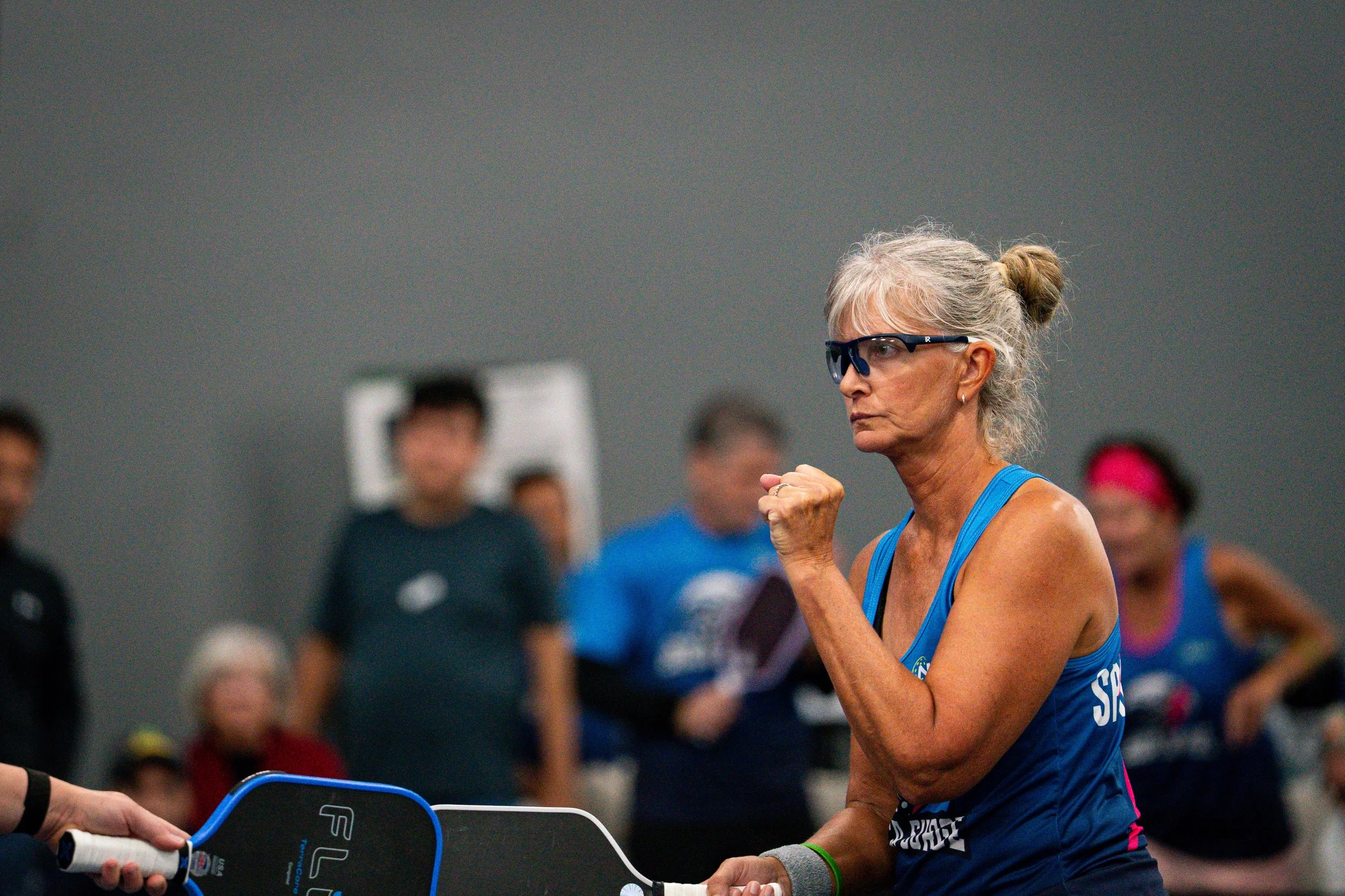 An older woman with gray hair tied in a bun, wearing glasses and a blue sports tank top, is participating in a fitness event, punching a pad held by another person with a blurred group of spectators in the background.