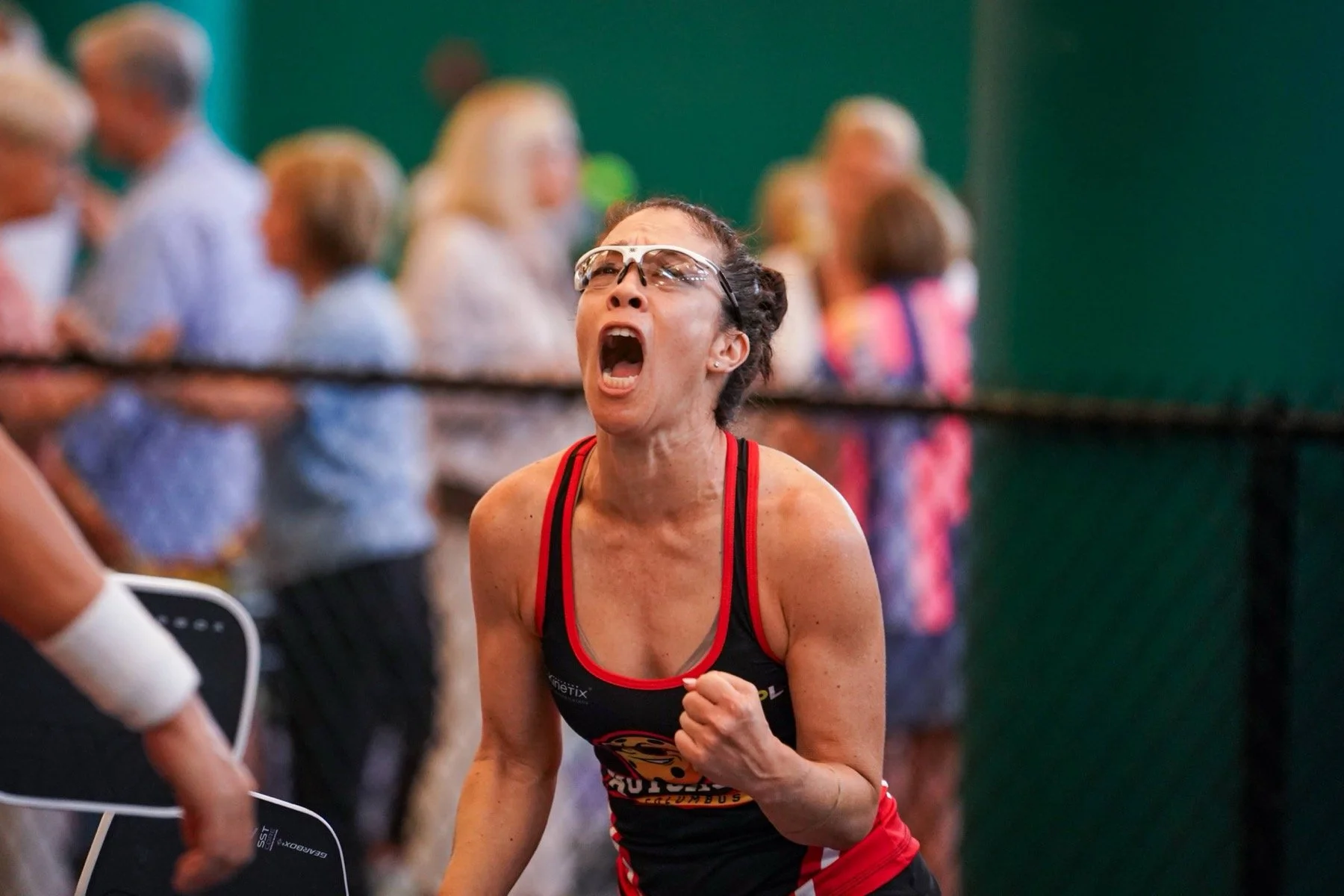 An woman with glasses wearing a black and red athletic tank top is running, with her mouth open and clenched fist, in a sports event. There are blurred people in the background.
