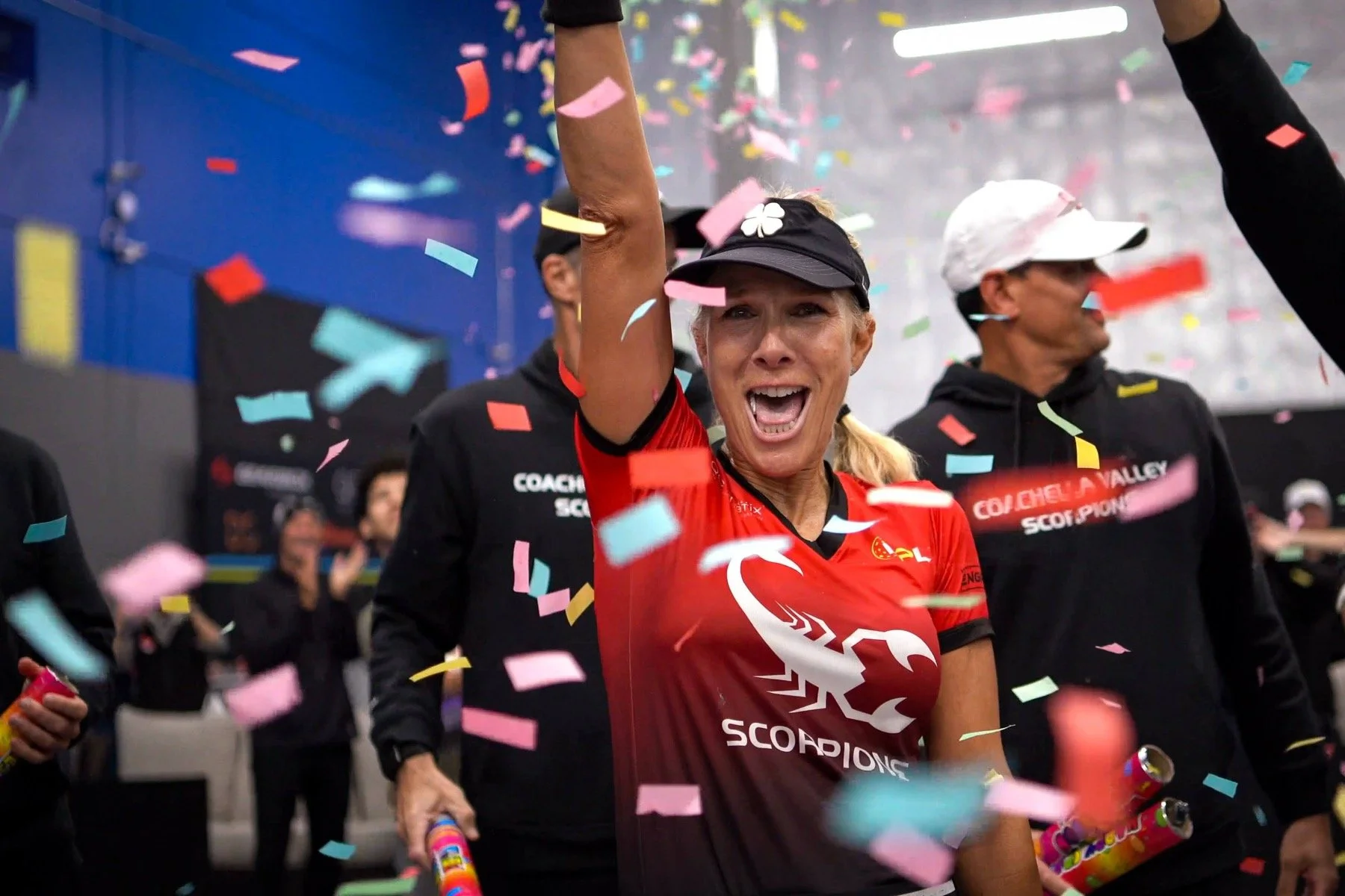 A woman celebrating with a joyful expression, surrounded by colorful confetti, wearing a red sports shirt and a black cap, holding party poppers.