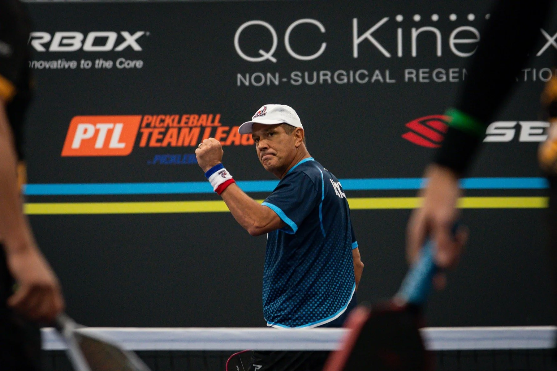 A man in a blue sports shirt and white cap clenches his fist in a victorious gesture during a pickleball match, with a black background displaying the logos for RBOX, QC Kínē non-surgical regeneration, and the PTL Pickleball Team League.