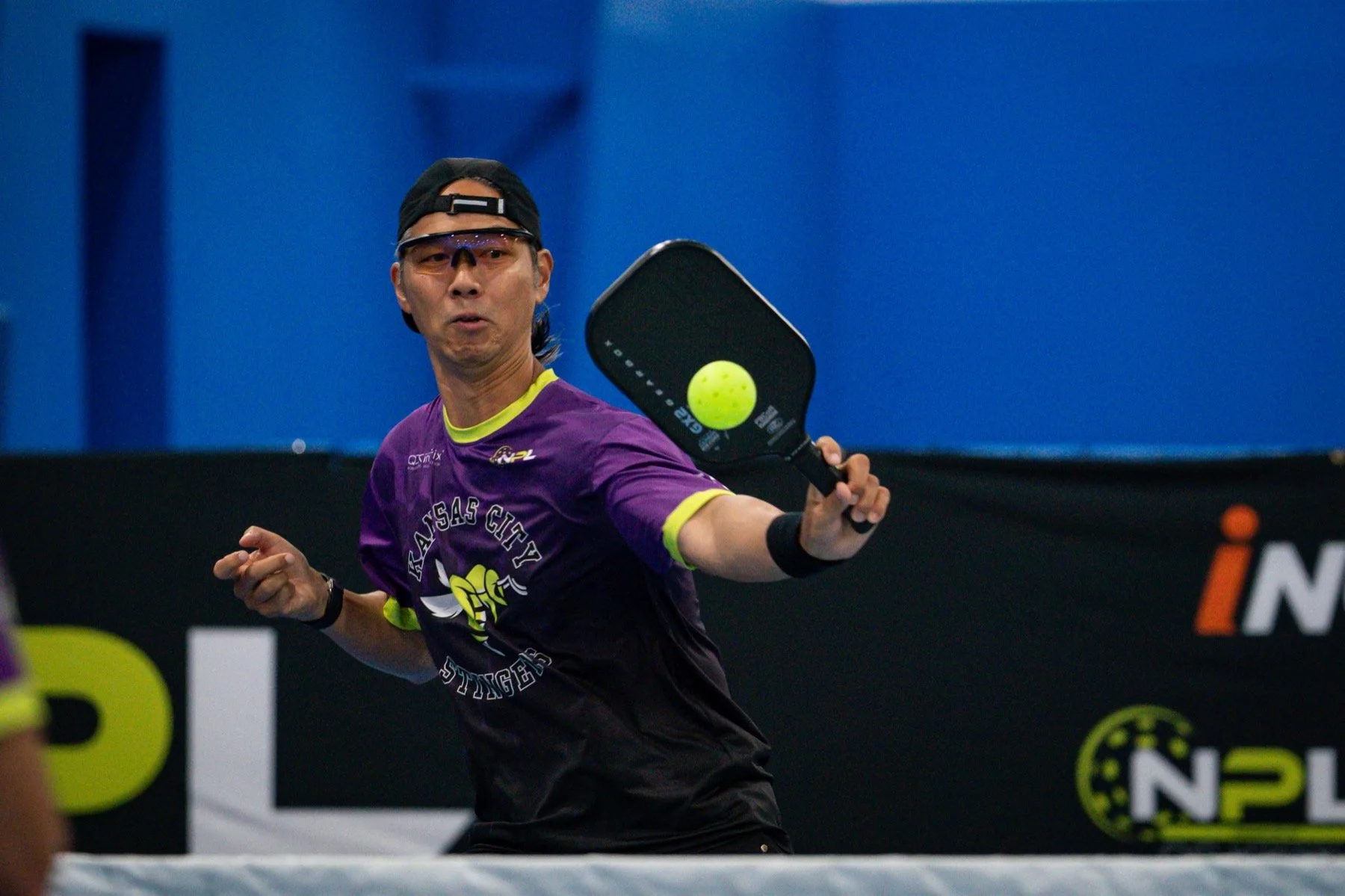 A male pickleball player in a purple Kansas City Stingers jersey, wearing protective glasses and a black cap, is hitting a yellow pickleball during a match.