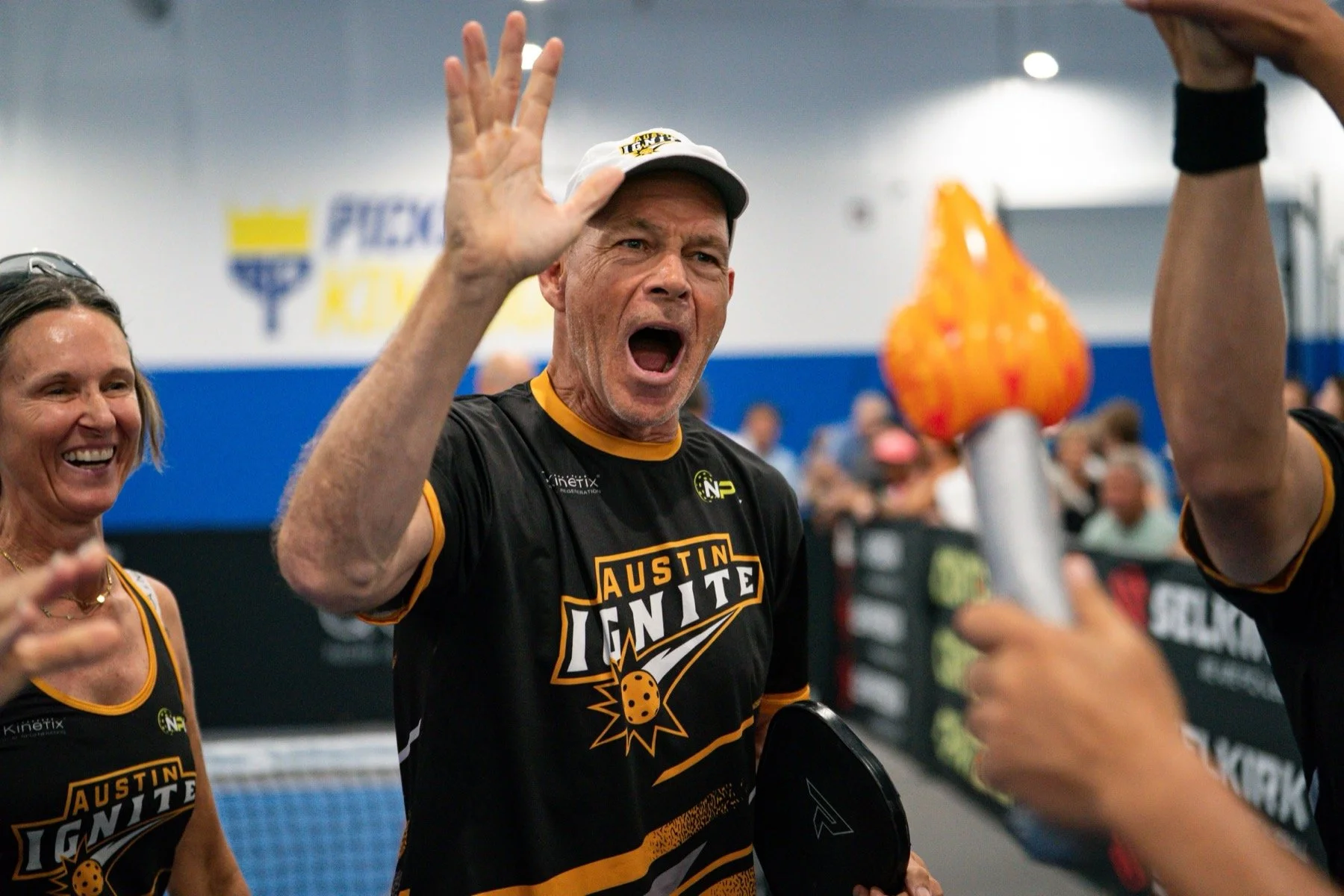 A man wearing a black and yellow Austin Ignite sports jersey and a white cap waves his hand while celebrating, surrounded by people at an indoor sports event.