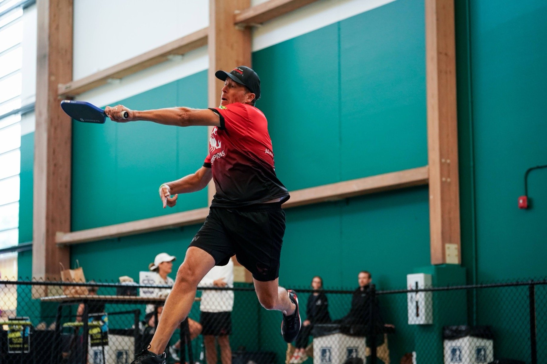A man playing pickleball indoors, wearing a red and black sports shirt, black shorts, a black cap, and holding a pickleball paddle.