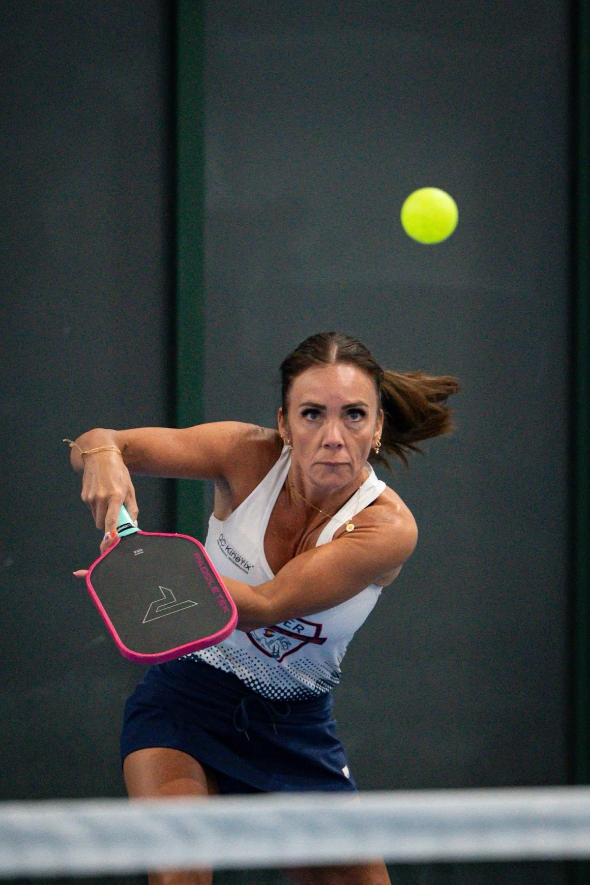 A woman playing pickleball, preparing to hit a green ball with a paddle, wearing a white tank top and navy shorts.