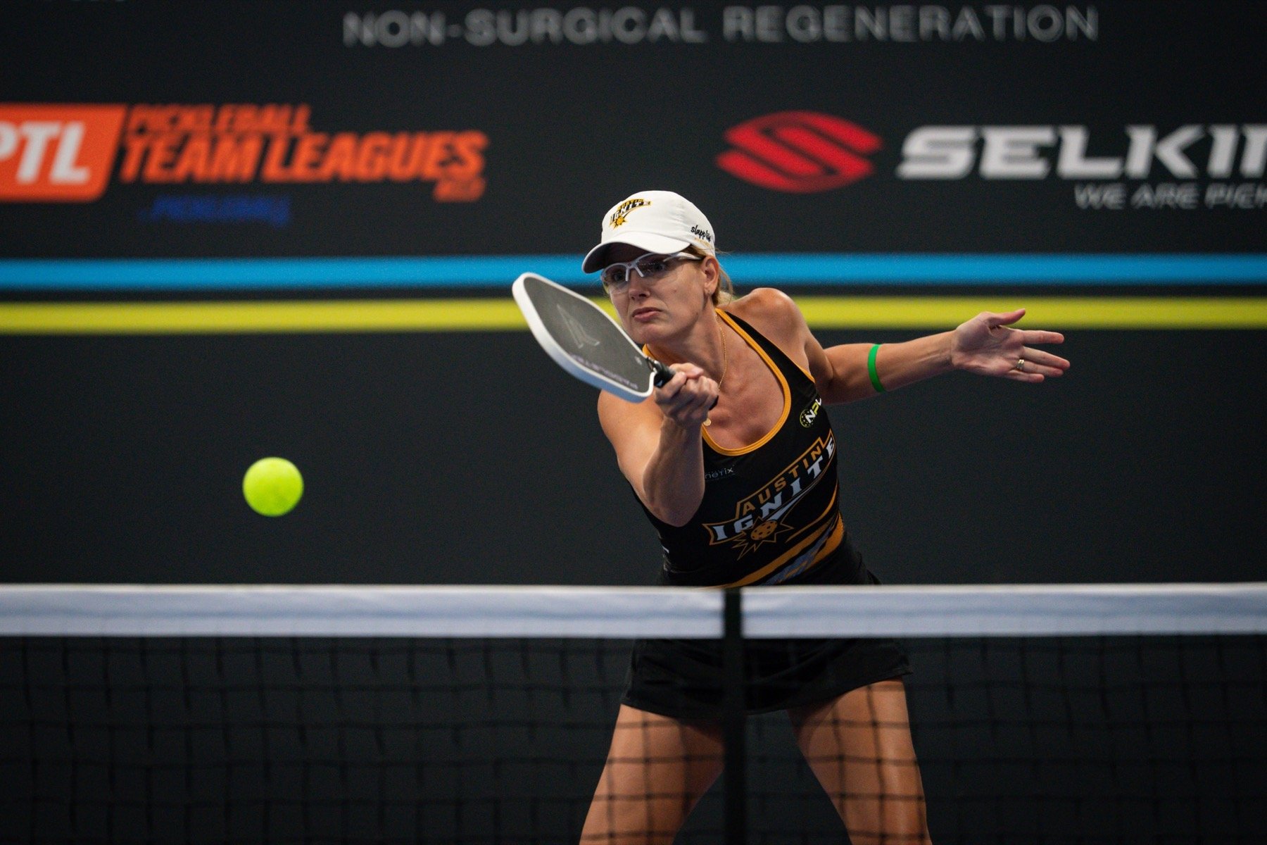 A woman wearing a white cap, sunglasses, and a black and yellow sports outfit with the word 'AUSTIN' on it, is playing pickleball.
