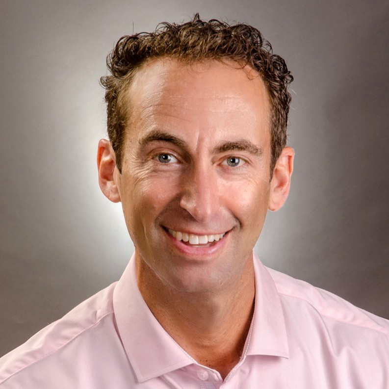 A smiling man in a pink dress shirt with short, curly brown hair, against a neutral gray background.