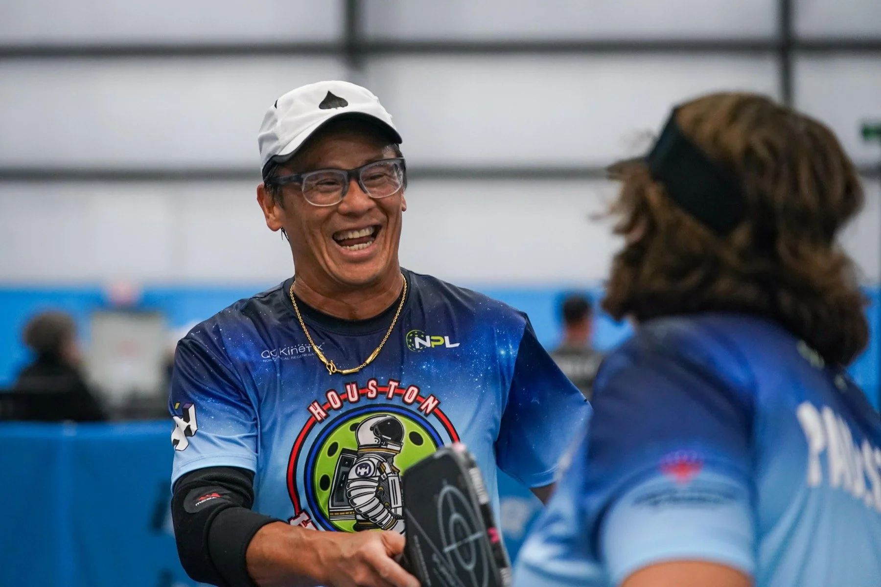 A man wearing glasses, a white cap with a spade symbol, and a Houston Rockettes jersey, smiling and holding a paddle, engaging with a woman in blue sportswear inside an indoor sports facility.