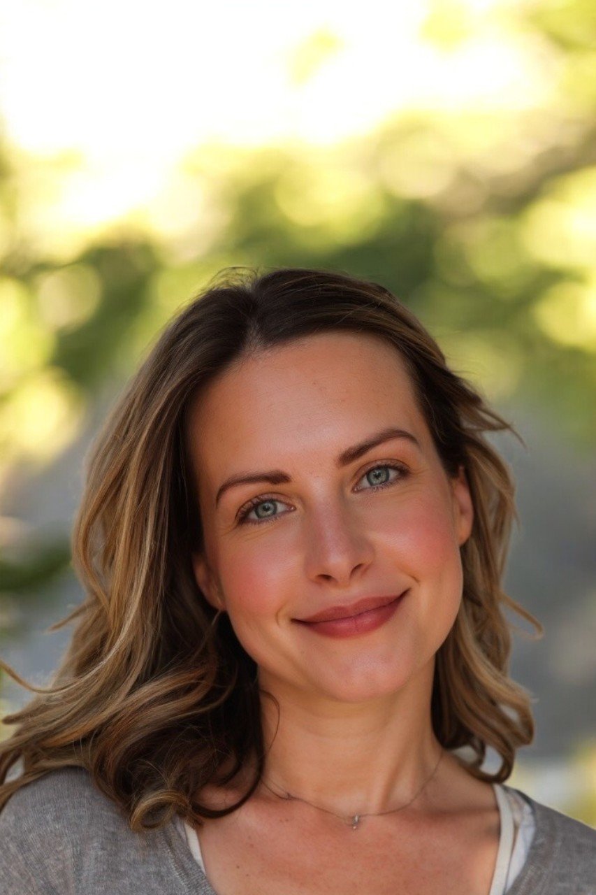 A woman with wavy brown hair and blue eyes smiling outdoors with a blurred green and yellow background.
