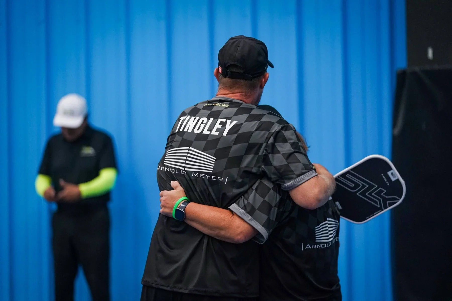 Two men hugging in front of a blue wall, one holding a tennis racket, the other wearing a black cap and shirt with 'TINGLEY' on the back.