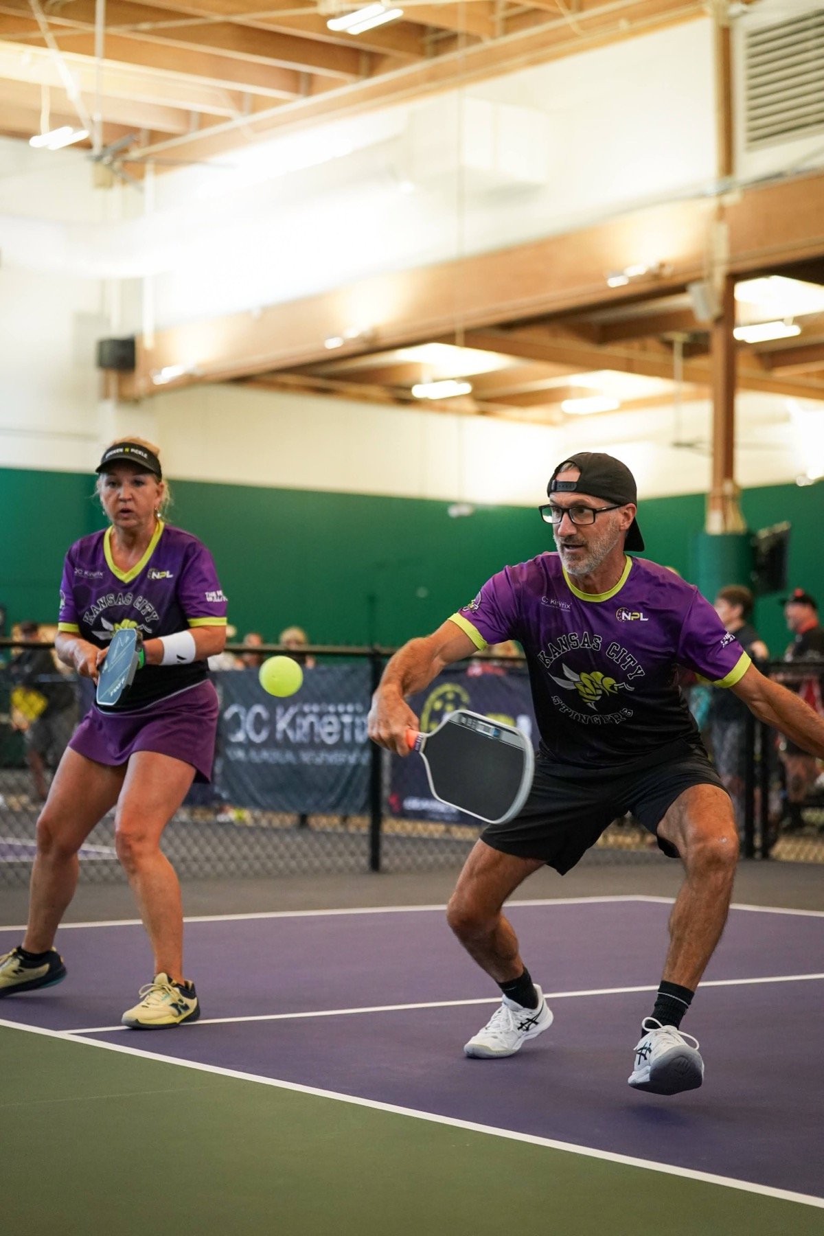 Two older adults playing pickleball on an indoor court, both wearing purple shirts with 'Kansas City Spinners' logo, one woman holding a paddle and the man reaching to hit a ball with a paddle.