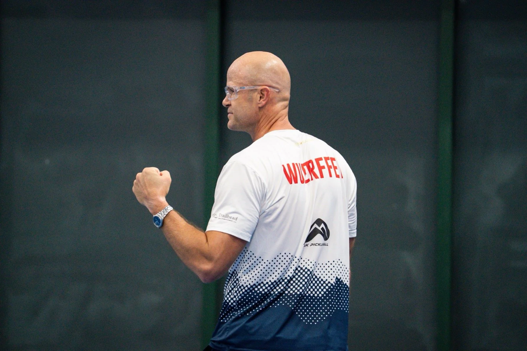 A bald man wearing glasses and a white sports shirt with red and black text, celebrating with a clenched fist during a sports event against a dark background.