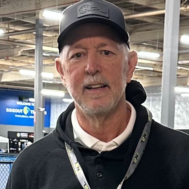 Man with a beard and mustache wearing a baseball cap, black jacket, and white polo shirt, standing indoors.