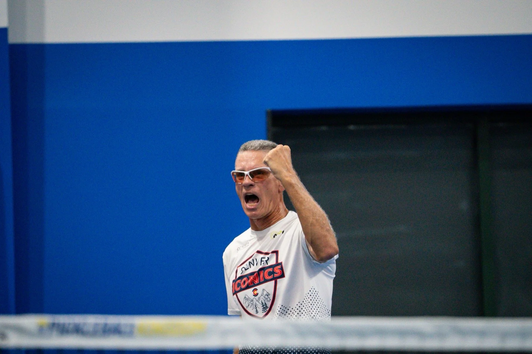 Man celebrating intensely, wearing sports sunglasses and a white Denver Iconics jersey, with fists clenched and mouth open, inside a sports facility with a blue wall background.