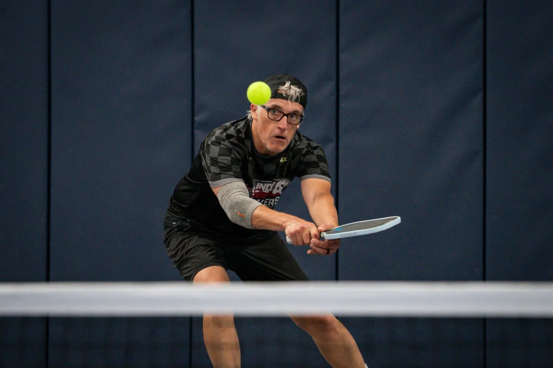 A man wearing glasses, a black cap backwards, a black sports shirt, and shorts playing pickleball indoors. He is lunging forward with his arm extended, holding a tennis racket, about to hit a pickleball.