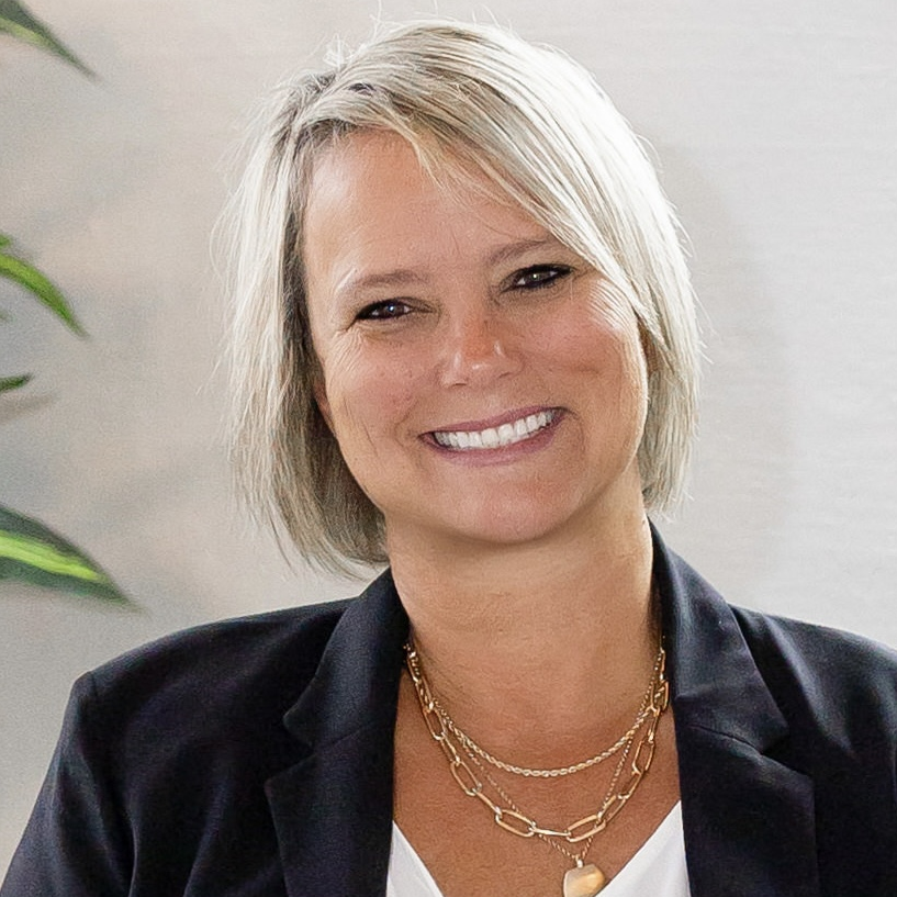 A woman with short blonde hair, smiling, wearing a black blazer and layered gold necklaces, seated indoors with a light-colored wall and green plant in the background.