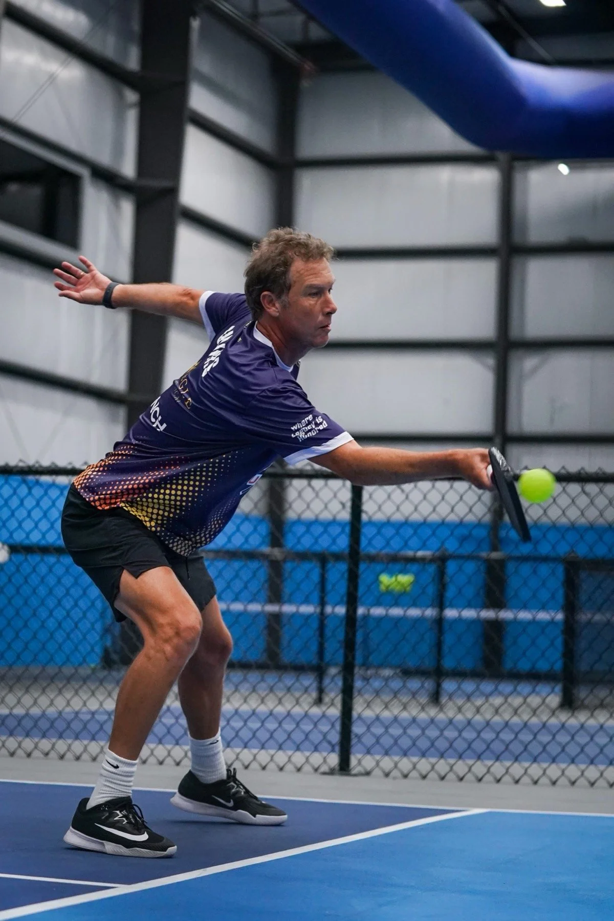 A man playing pickleball inside a sports facility, preparing to hit a bright green ball with a paddle, wearing a dark sports shirt, shorts, and athletic shoes.