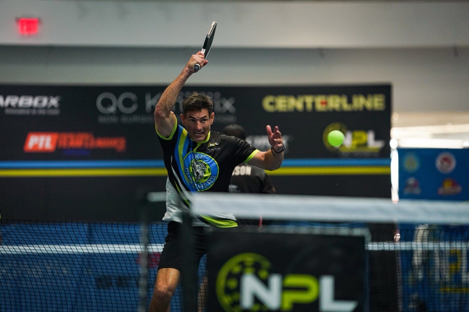 A man playing table tennis indoors, holding a paddle in his right hand while hitting a yellow tennis ball. He is wearing a black shirt with blue, green, and white accents, and has a focused or intense expression on his face. The background features advertising banners and a table tennis net.