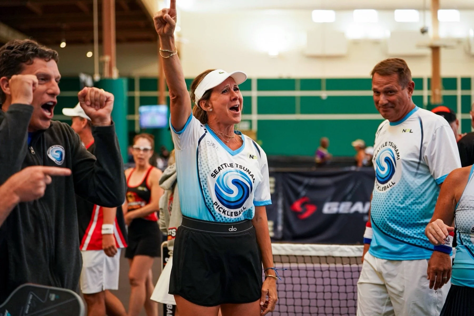 Women and men at an indoor pickleball court, celebrating and talking after a game, with a woman raising her hand in victory, wearing a Seattle Tsunami Pickleball shirt.