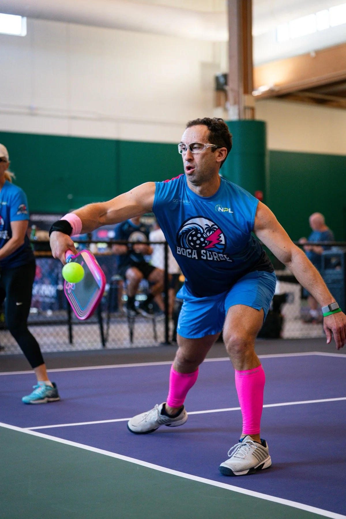 A man playing pickleball indoors, wearing a blue sports jersey with a logo and text, pink knee-high socks, white sneakers, and safety glasses, preparing to hit a neon green pickleball with a pickleball paddle.