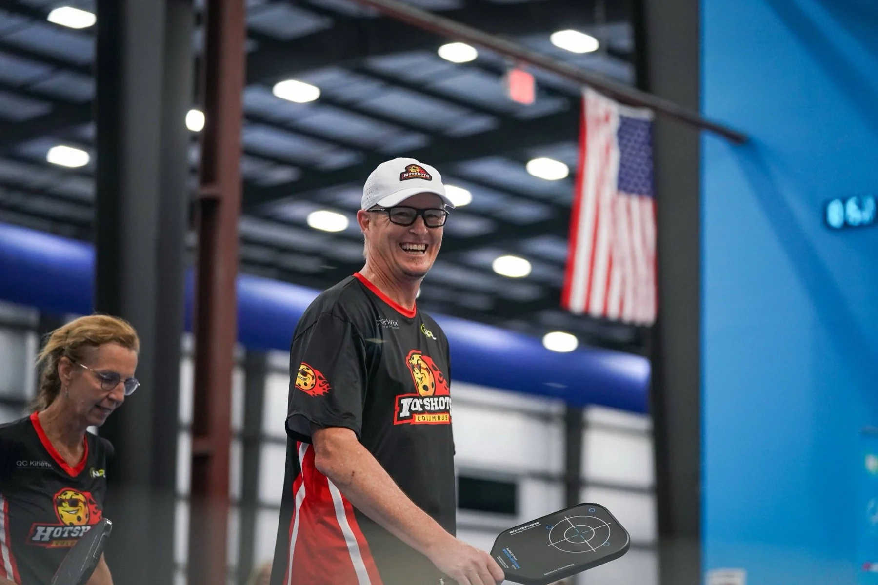 A man wearing glasses and a white cap is smiling and standing in an indoor arena. He is dressed in a black sports jersey with red and yellow logos, holding a pickleball paddle.