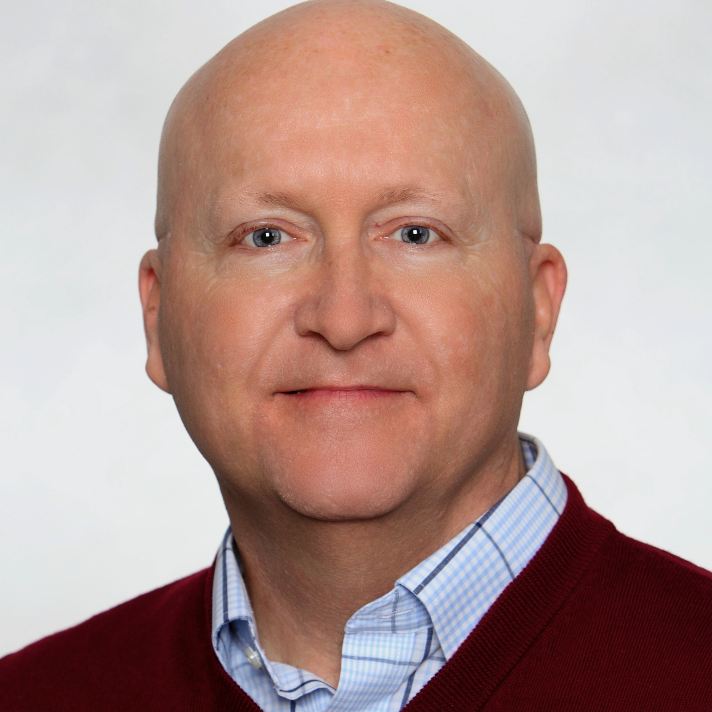 Close-up of a man wearing a blue and white checkered shirt and a red sweater, with earbuds visible around his neck.