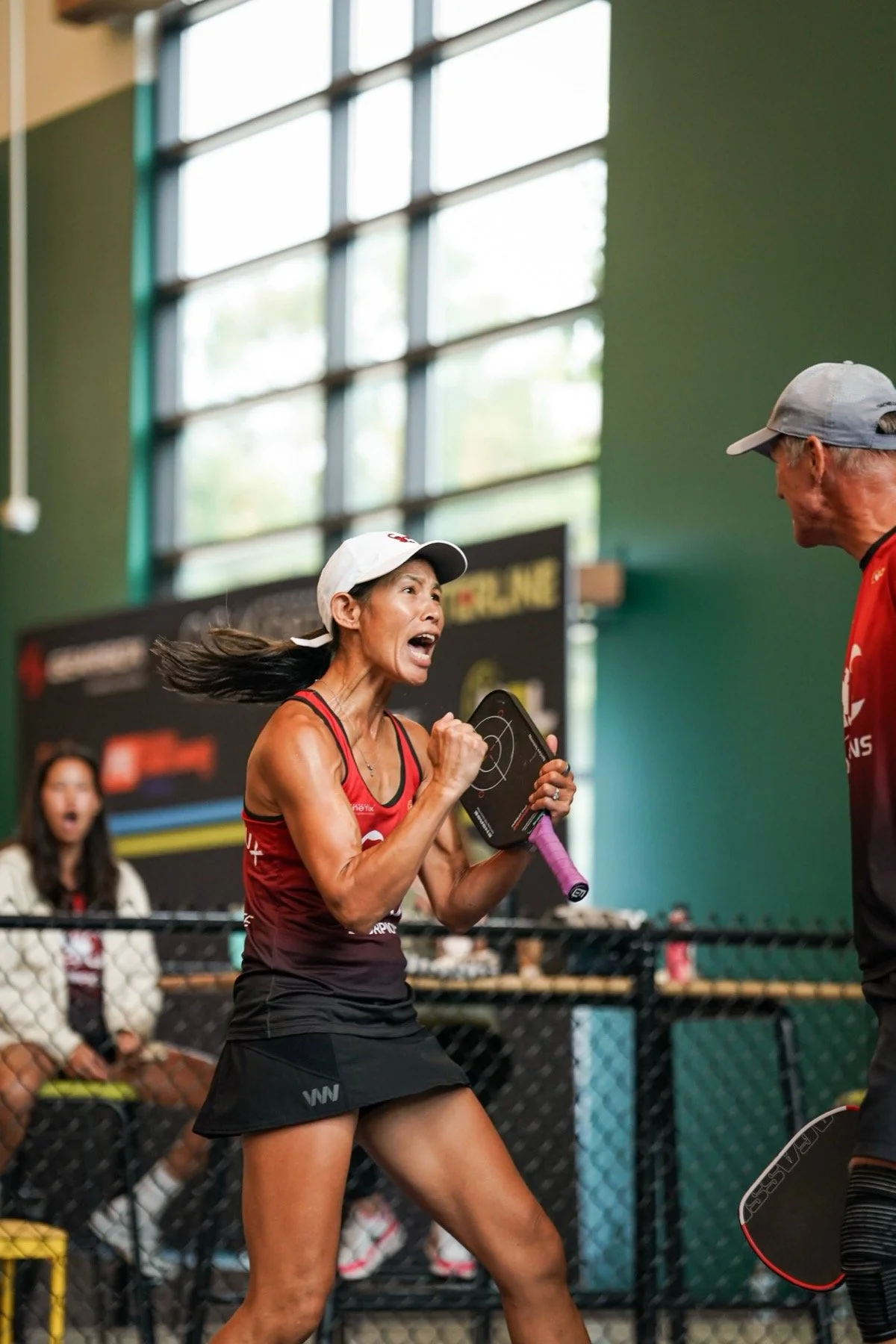 Female athlete celebrating during a sporting event in an indoor gym, wearing a red sports uniform and white cap.