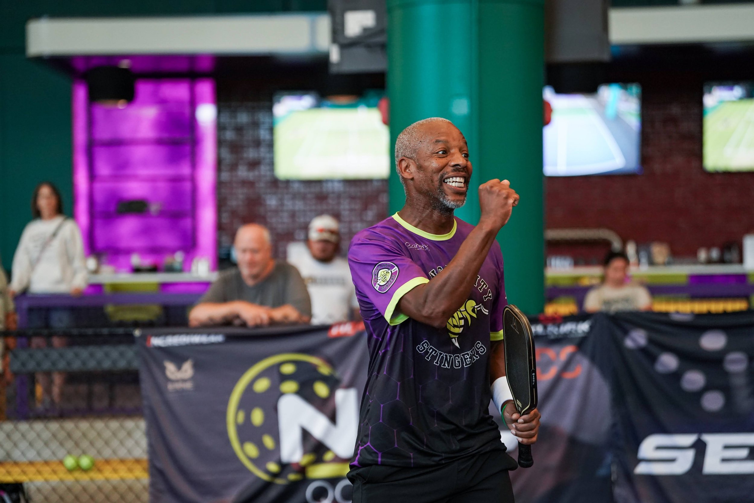 A man with gray hair and beard smiling and holding a ping pong paddle, wearing a purple and black sports shirt in an indoor sports facility with multiple TV screens and people in the background.