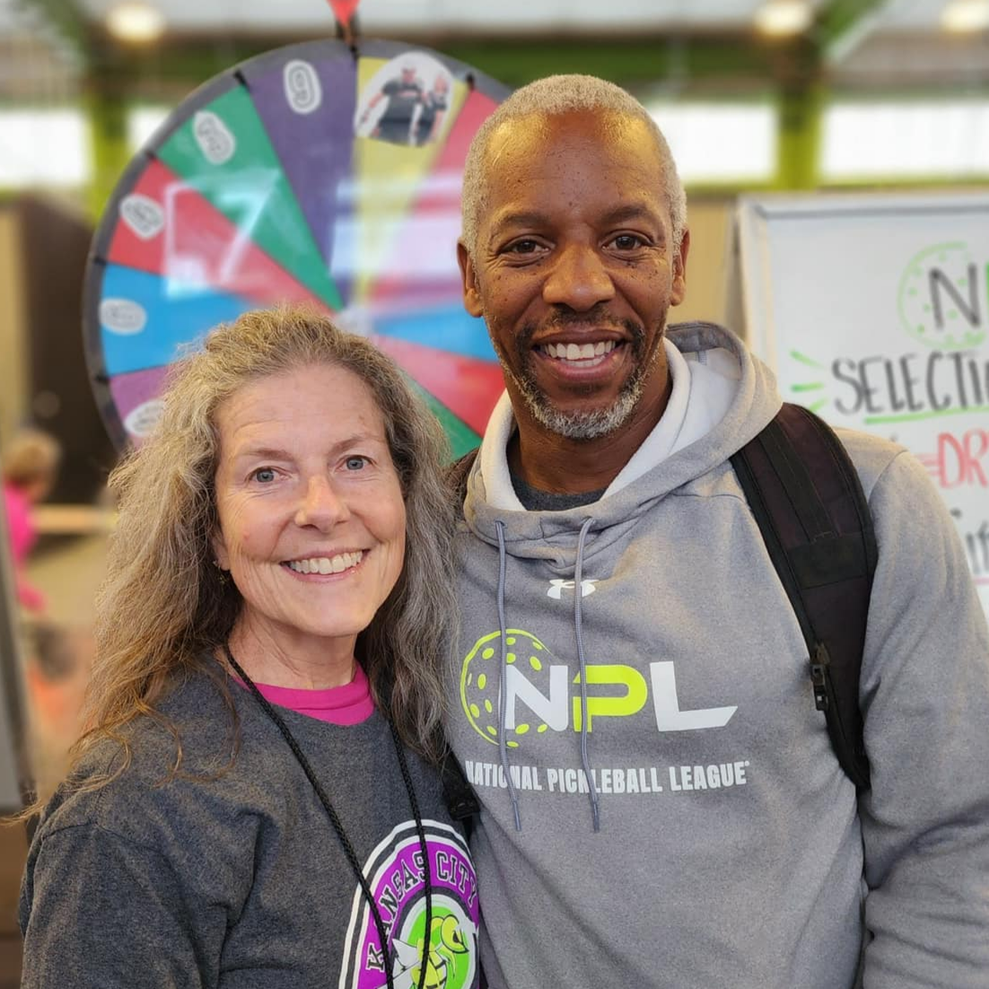Two smiling people, a woman with long gray hair and a man with short gray hair, standing together at an event with a colorful spinning wheel in the background. The woman wears a gray shirt with a purple and yellow logo, and the man wears a light gray hoodie