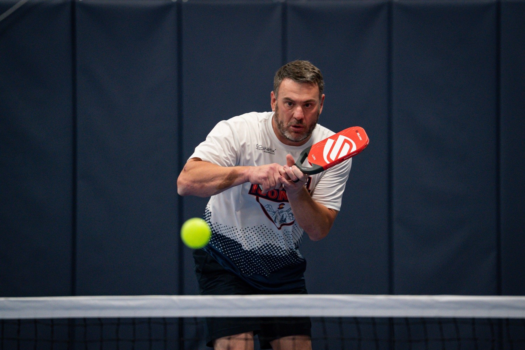 Man playing pickleball, preparing to hit a neon green ball with a red pickleball paddle, indoors against a dark wall.