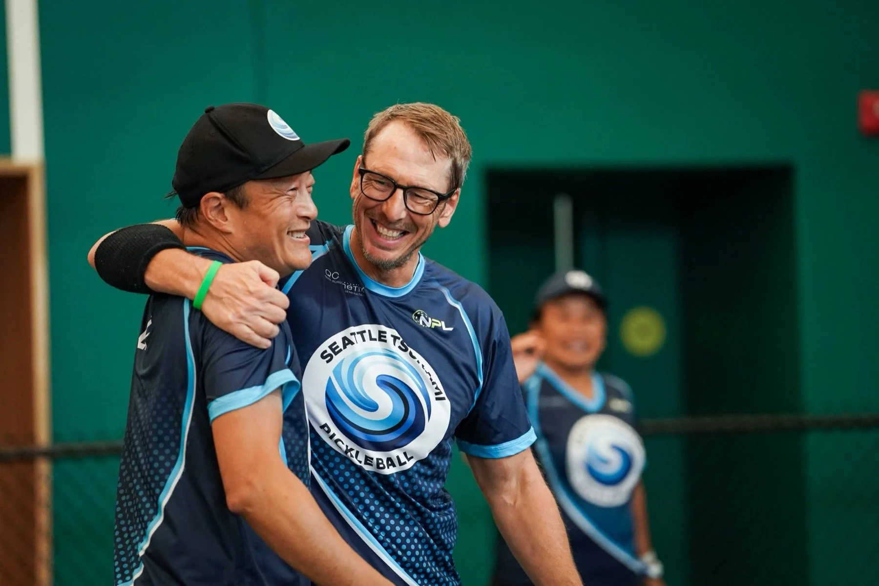 Two men in matching blue Seattle Tsunami Pickleball jerseys sharing a joyful moment with one patting the other's shoulder and both smiling.