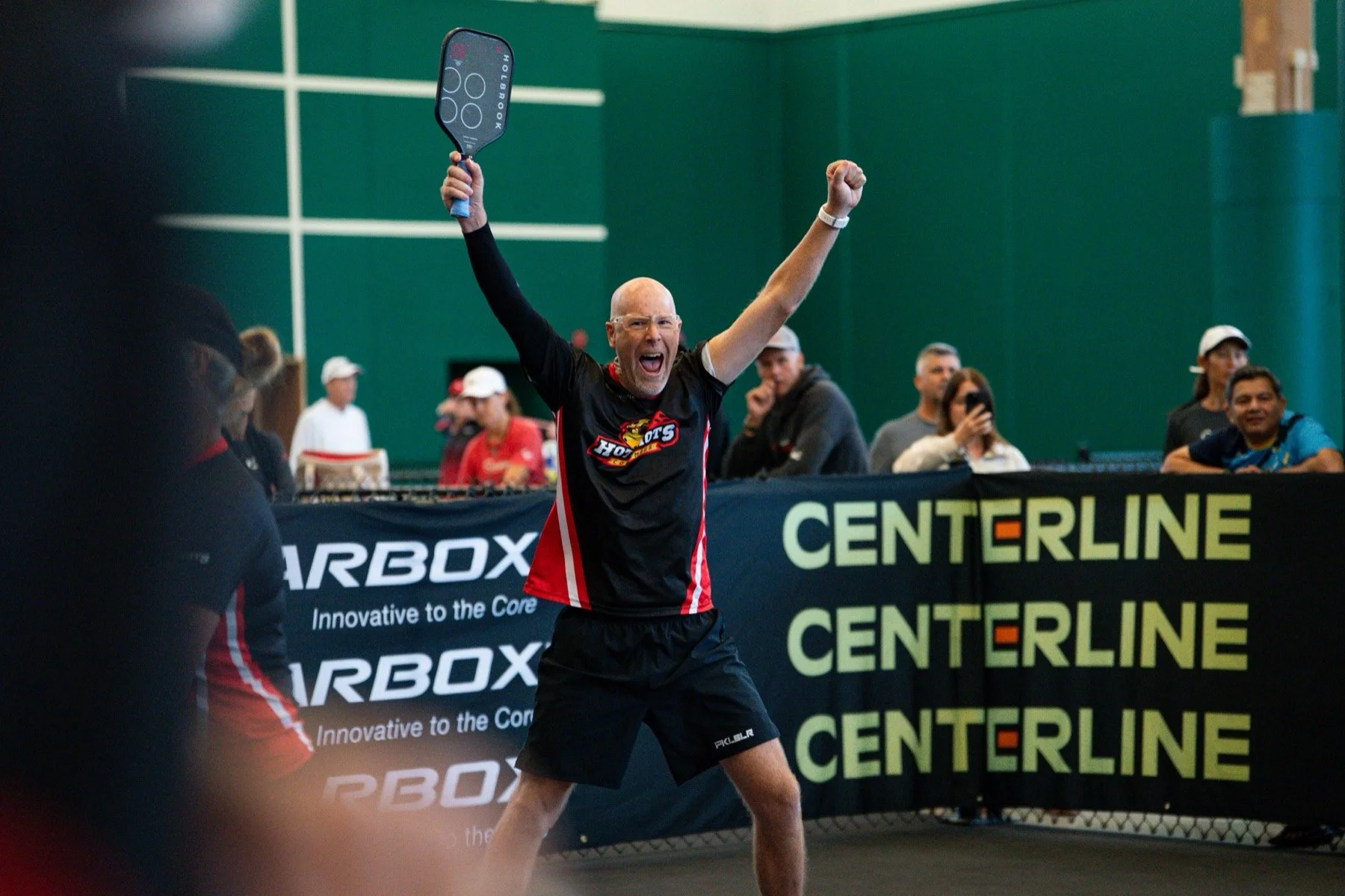 A man in a black and red sports shirt celebrating with arms raised on a tennis court, holding a paddle in his right hand. Behind him, there are spectators and a banner with the words 'CENTERLINE'. The background features a green wall and more spectators observing the event.