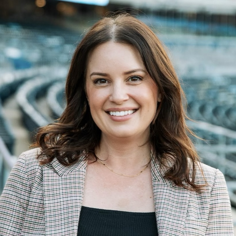 A smiling woman with shoulder-length brown hair, wearing a plaid blazer over a black top, outdoors with stadium seating in the background.