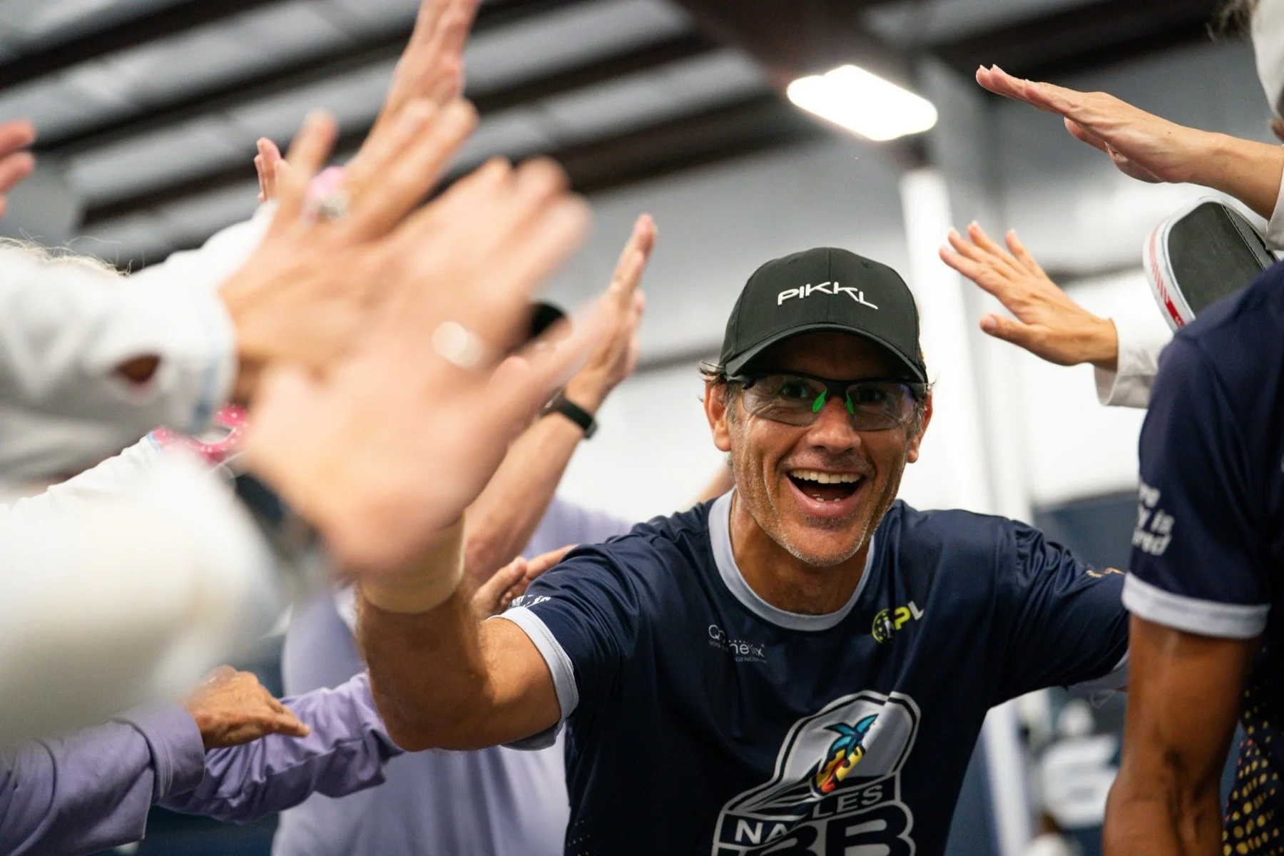 A man wearing a black cap with the word 'PIKKL', safety glasses, and a navy cycling jersey is smiling and celebrating. He is surrounded by people extending their hands towards him in a gesture of celebration or encouragement. The scene appears to be indoors, likely at a sporting event or team gathering.