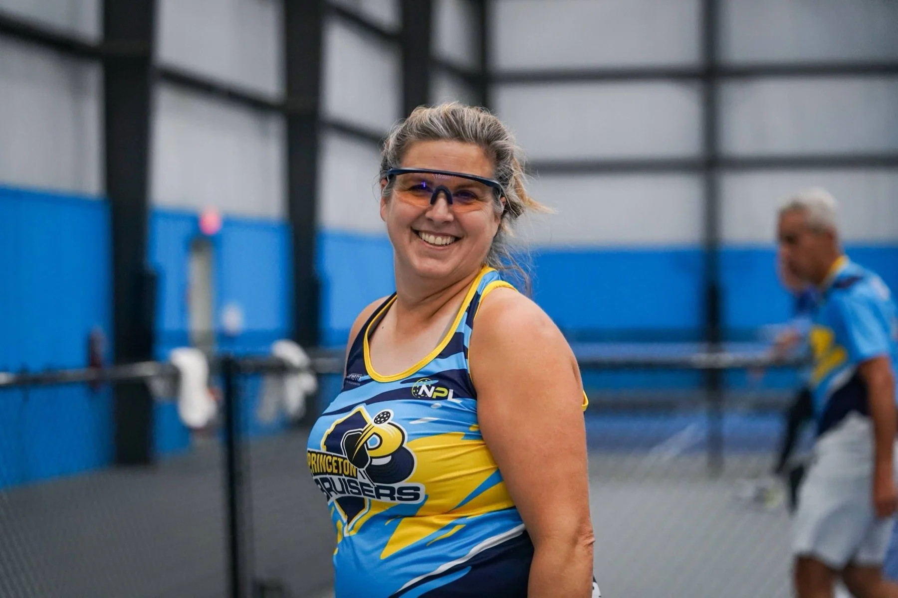 A smiling woman with glasses wearing a blue and yellow sports jersey, standing inside a pickleball facility and another person blurred in the background.