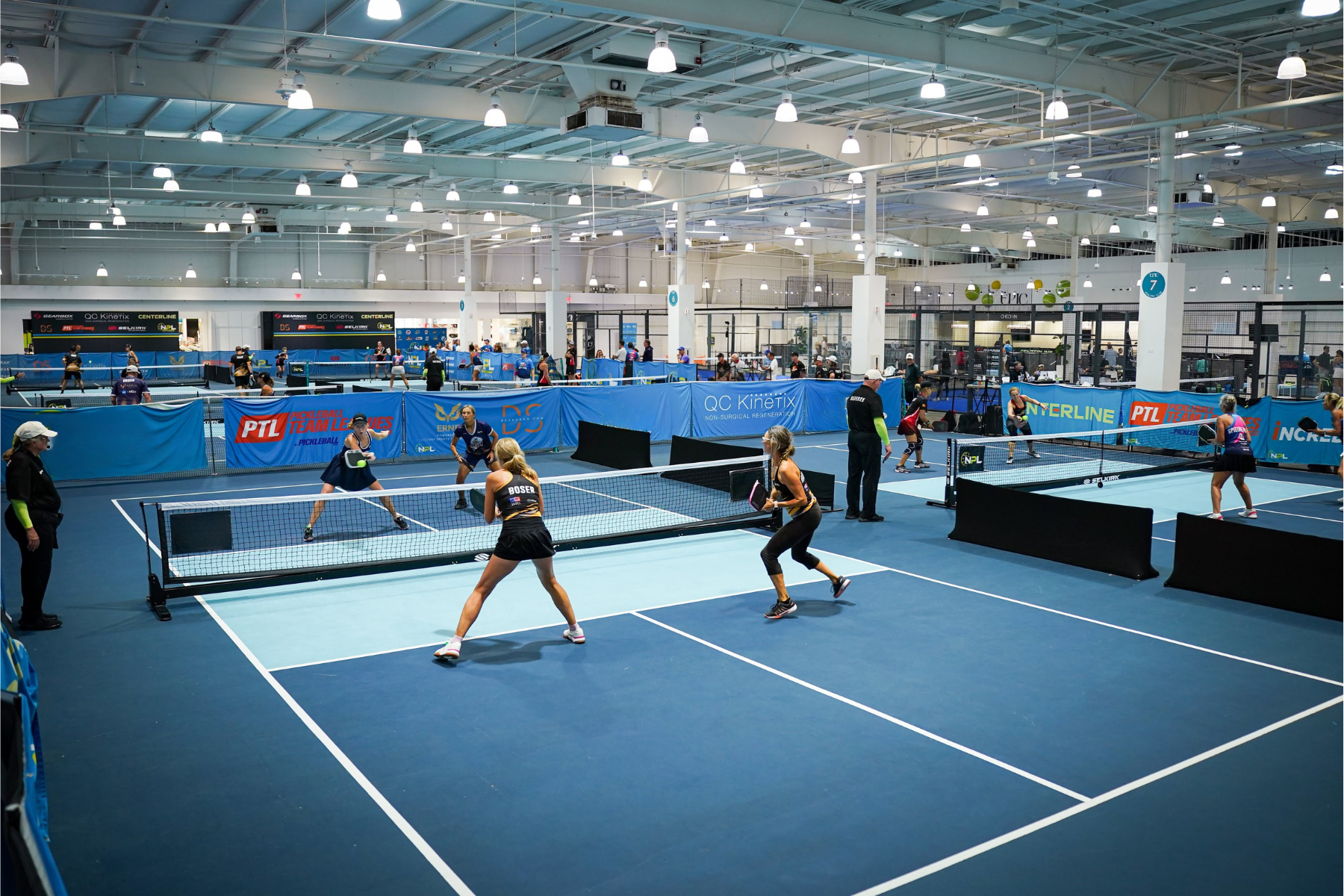 Indoor pickleball courts with players engaged in a doubles match. The courts are blue with white lines, surrounded by blue and green barriers and banners. The ceiling has numerous bright lights, and there are spectators and officials present.
